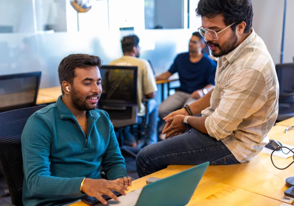 Two coworkers talking and working at a laptop in an open office, with colleagues in the background