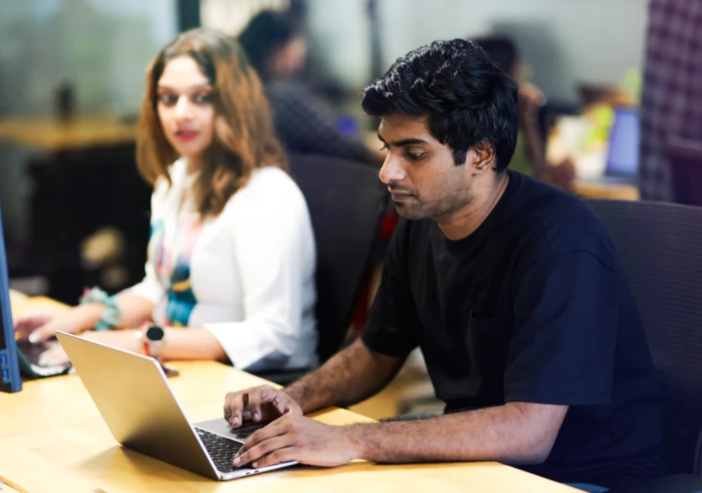 Man working on a laptop at a desk in an office, with a colleague in the background.