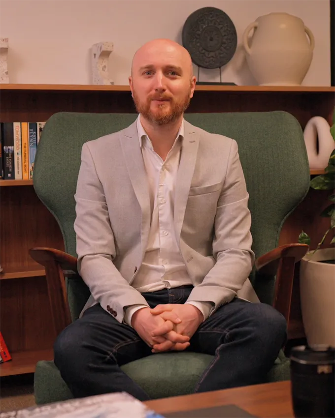 A man in a light jacket sitting in a green armchair in front of a bookshelf with decorations.