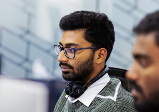 Employee wearing glasses and a headset working at a computer in an office, with a coworker blurred in the foreground.