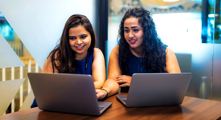 Two women working together on laptops at a table in a modern office